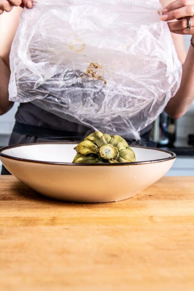 White bowl with microwaved artichoke inside of it. The chef is removing the plastic wrap from the bowl.