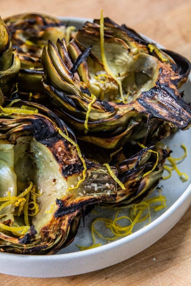 Grilled artichoke halves set in a grey dish topped with lemon peel curls.