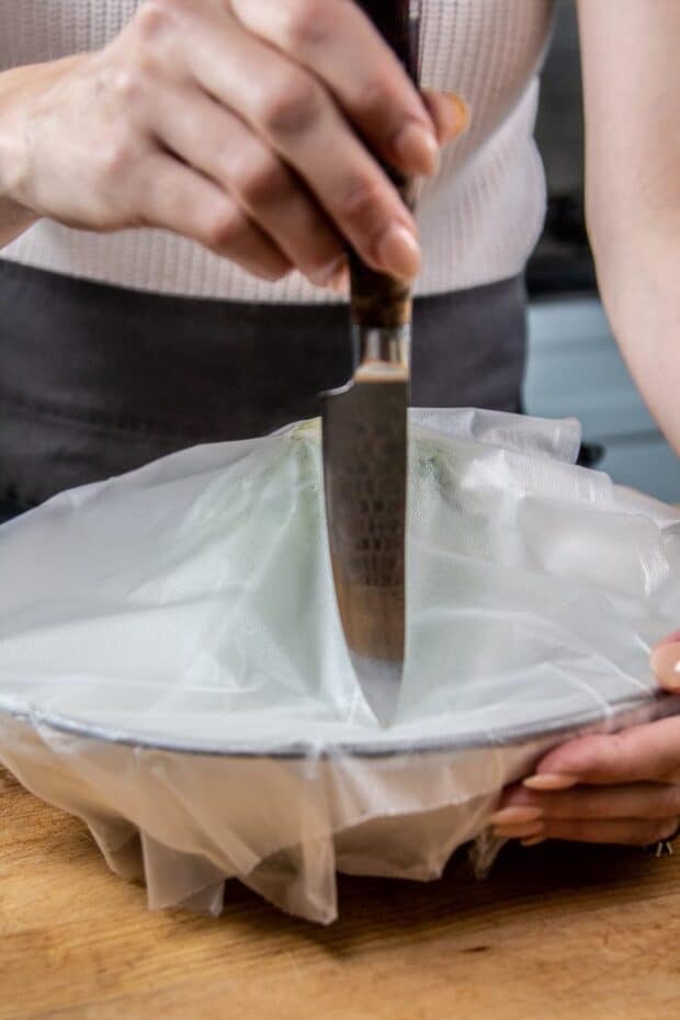 A microwave-safe bowl  with an artichoke inside. The bowl is covered with a press n' seal wrap. The chef is ventilating the bowl by poking holes in the wrap with a knife. 