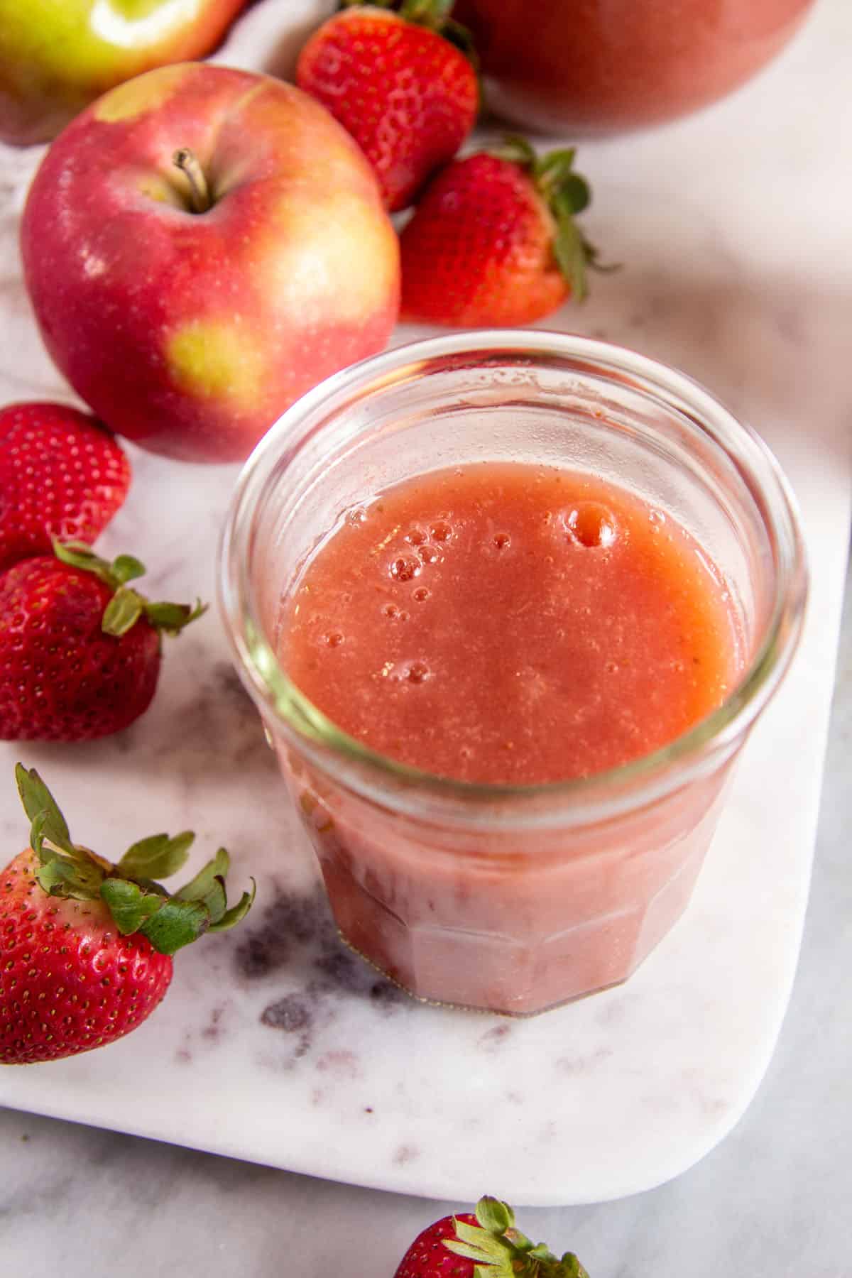 jar of strawberry applesauce on a marble background surrounded by apples & strawberries