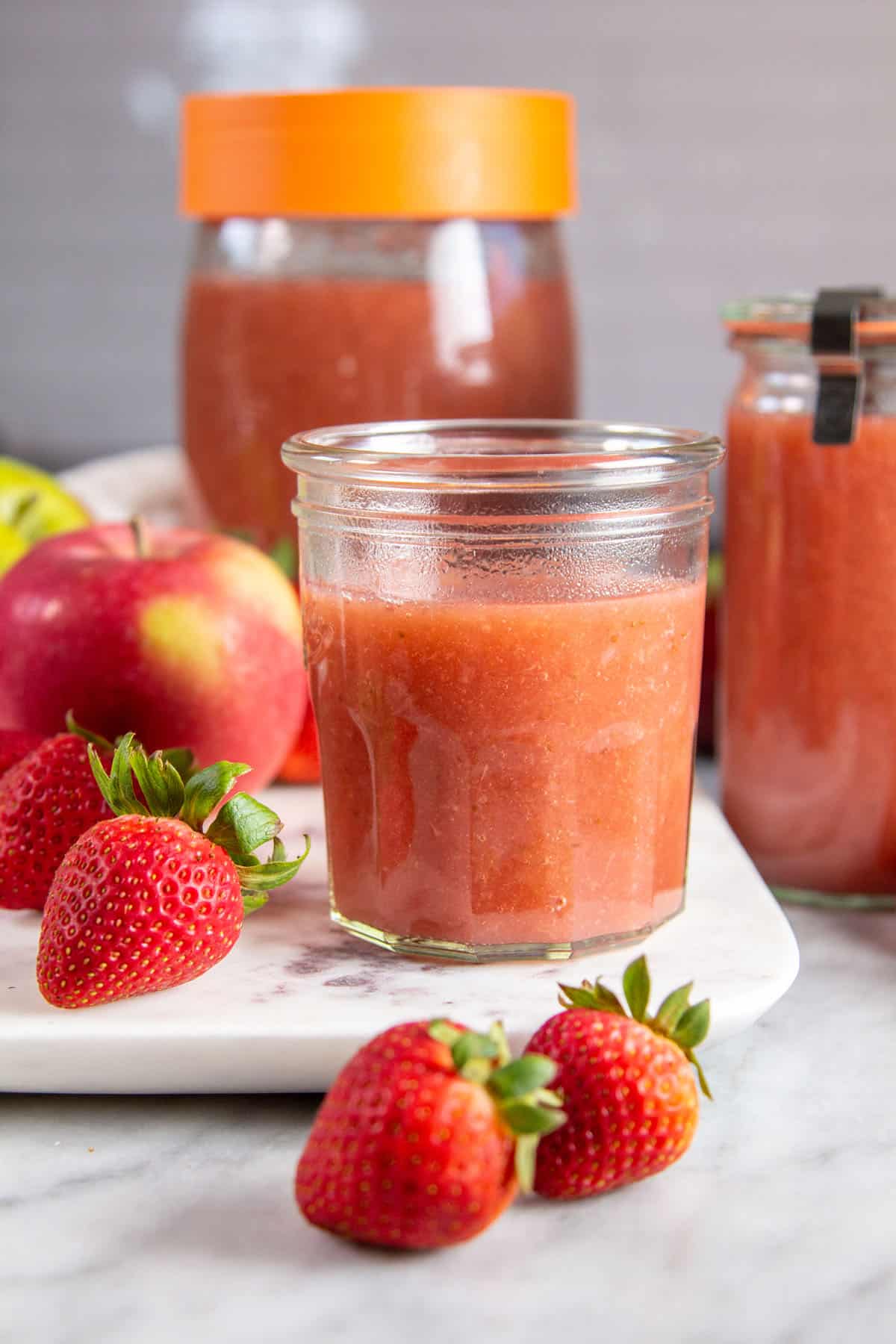 multiple jars of strawberry applesauce on a marble background surrounded by apples & strawberries