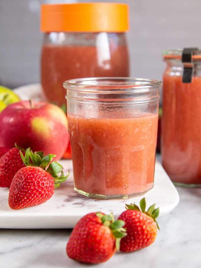 multiple jars of strawberry applesauce on a marble background surrounded by apples & strawberries