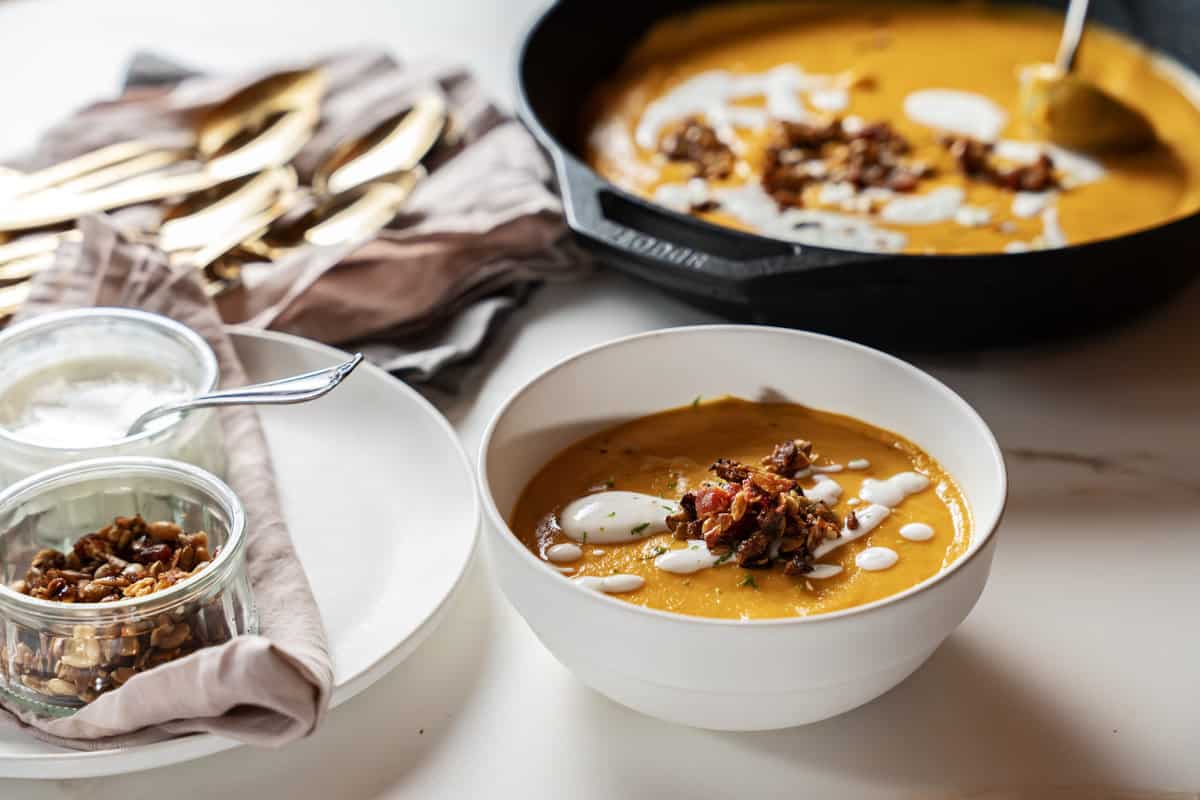 a bowl of roasted sweet potato soup garnished with lime yogurt and apple pepita bacon brittle next to a skillet full of the soup with a ladle in it. a pile of gold soup spoons on a napkin and small bowls with extra garnishes