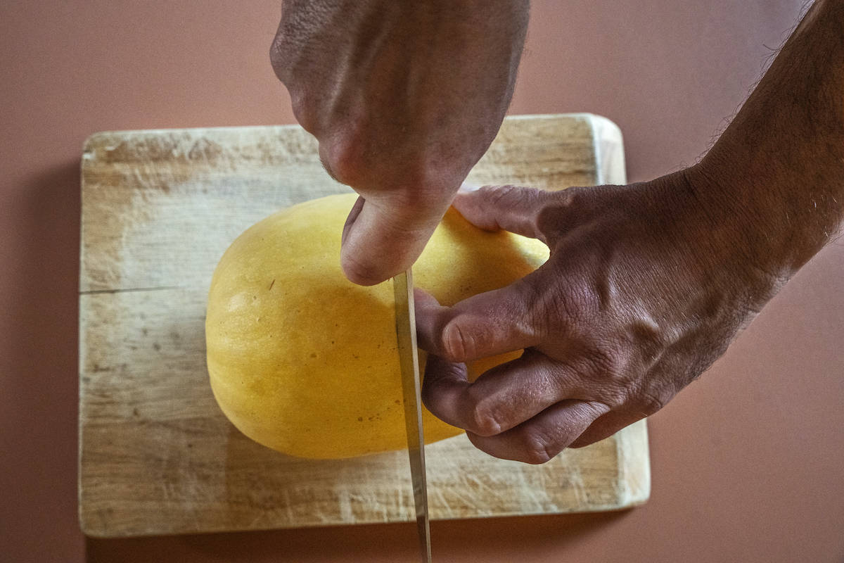 overhead view of two hands cutting a raw spaghetti squash in half horizontally on top of a cutting board