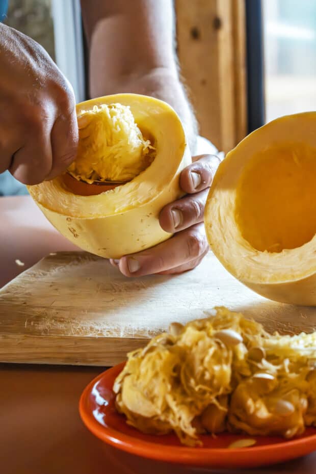 a spoon being used to scrape out the seeds and guts of a raw, halved spaghetti squash. one half is already scooped empty with the guts on a red plate.