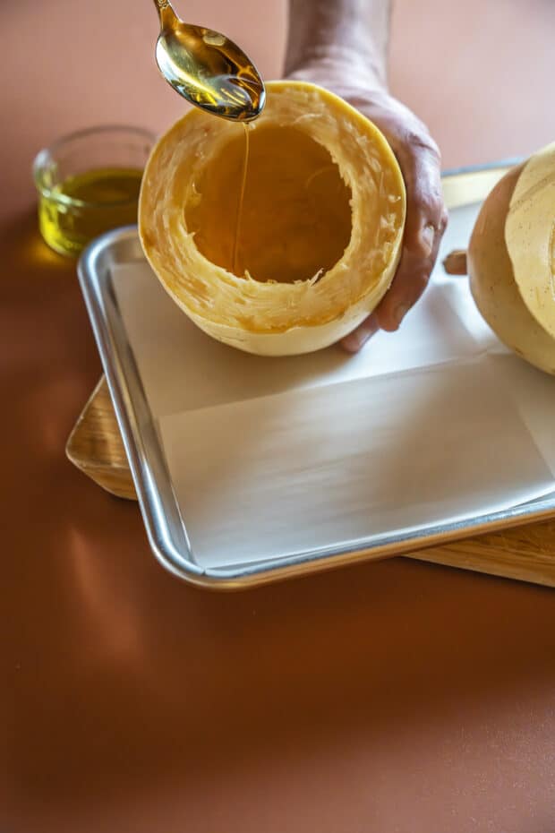 a spoon being used to drizzle olive oil on half of a raw spaghetti squash with seeds removed