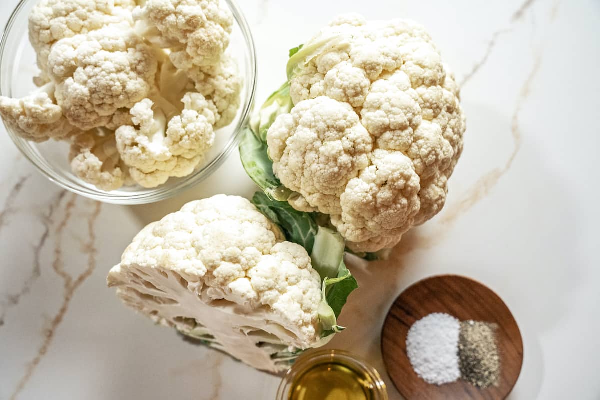 over head view of a whole head of cauliflower, a half head of cauliflower, cauliflower florets in a glass bowl, salt and pepper in a small brown bowl, oil in a small glass bowl to make cauliflower rice.