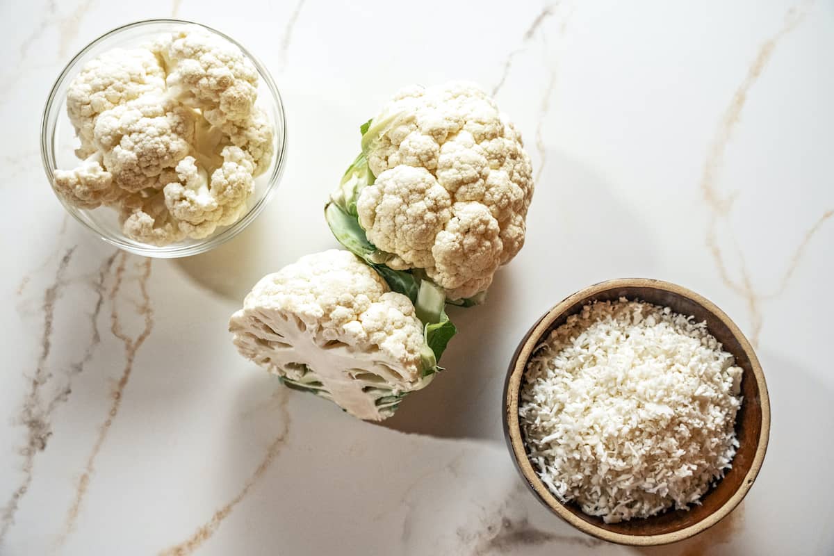 over head view of raw cauliflower florets in a glass bowl, raw whole cauliflower head, and a brown bowl full of raw grated cauliflower rice