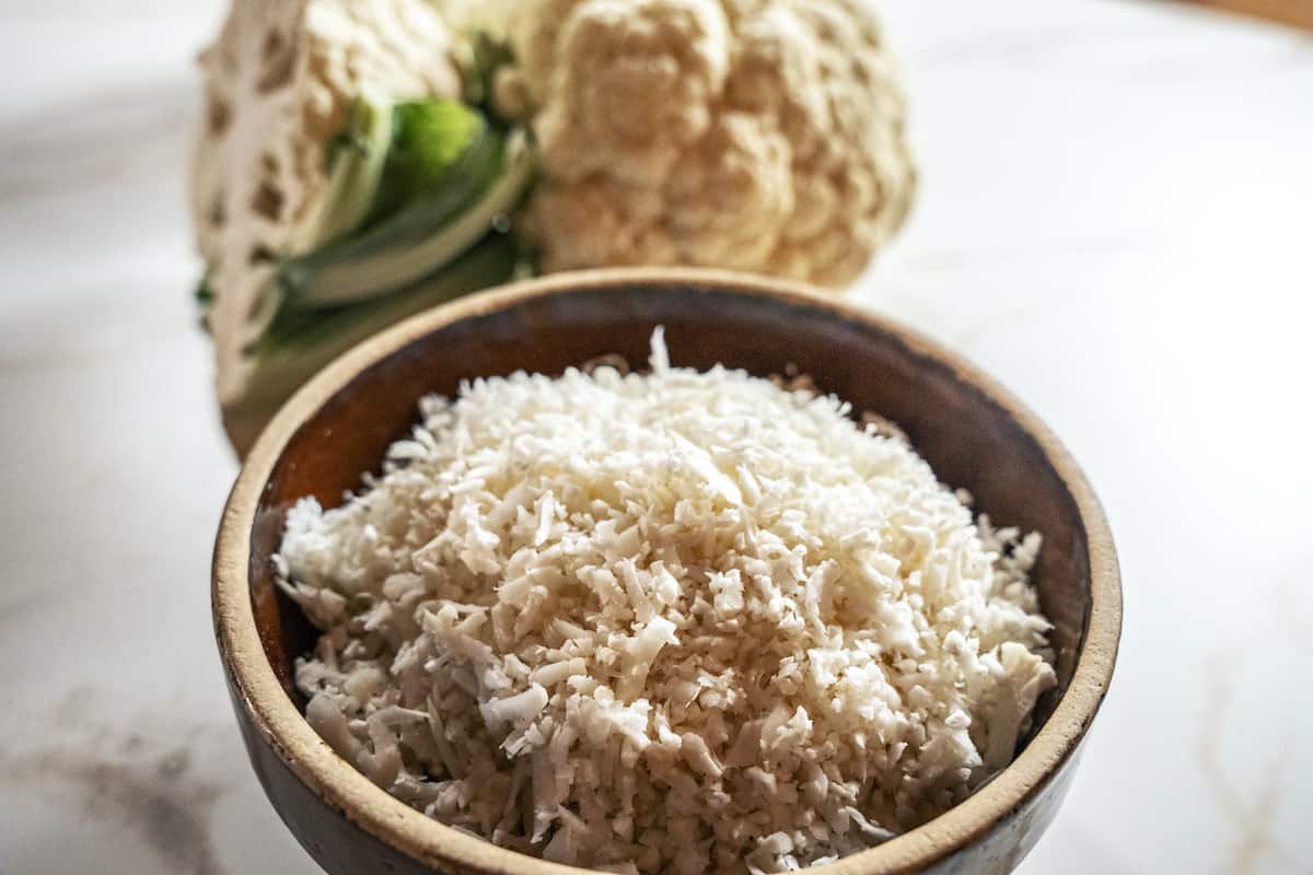 close up of raw grated cauliflower rice in a brown bowl with whole cauliflower heads in the background
