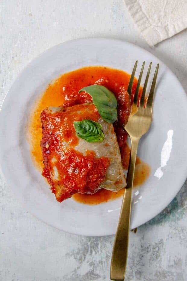 A cabbage roll on a serving plate with red pepper sauce and basil leaves. 