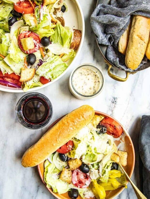 A single serving of salad in a bowl with a breadstick. A glass of wine, bottle of dressing, basket of breadsticks and larger serving bowl of salad is also in the shot.