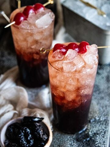 two glasses of homemade soda with cherrys in them next to a small bowl of prunes