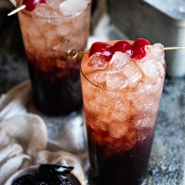 two glasses of homemade soda with cherrys in them next to a small bowl of prunes