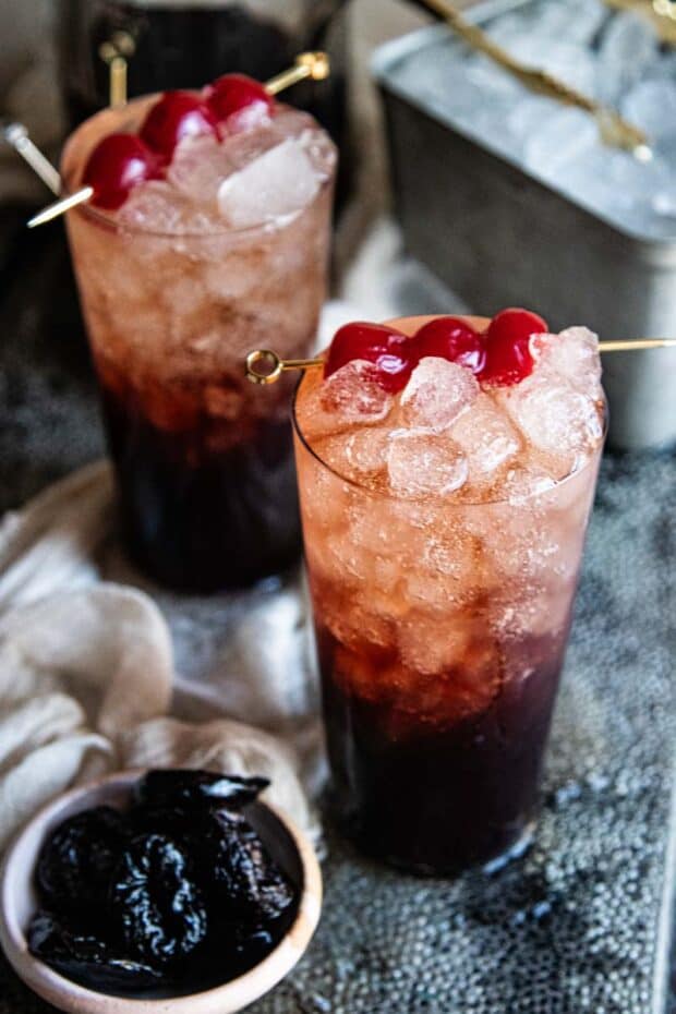 two glasses of homemade soda with cherrys in them next to a small bowl of prunes