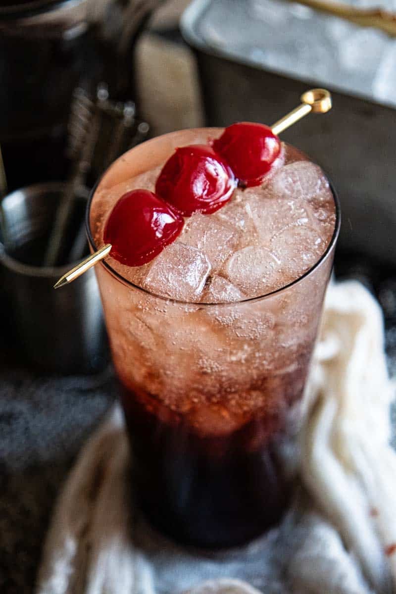 single glass of cherry dr. pepper with skewer on top with three cherries on it