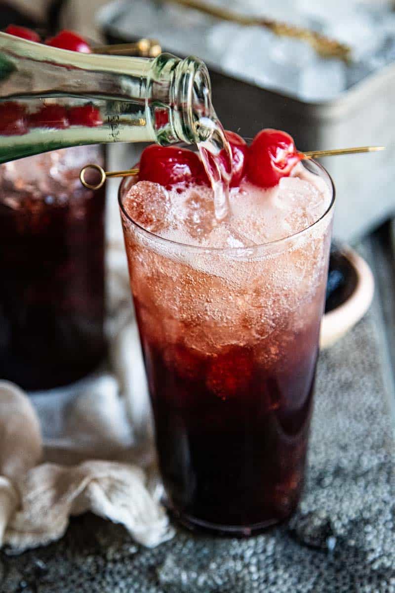 clear bottle of sparkling water pouring into a prepped glass with cherry dr. pepper syrup